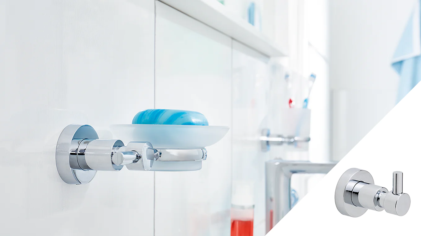 A chrome wall-mounted soap dish holds a blue bar of soap next to a white bathroom cabinet. In the corner, a separate chrome robe hook is displayed. Various toiletries are blurred in the background, and nearby, rolls of tesa tape are neatly stacked for convenient use. (This text has been generated by AI)