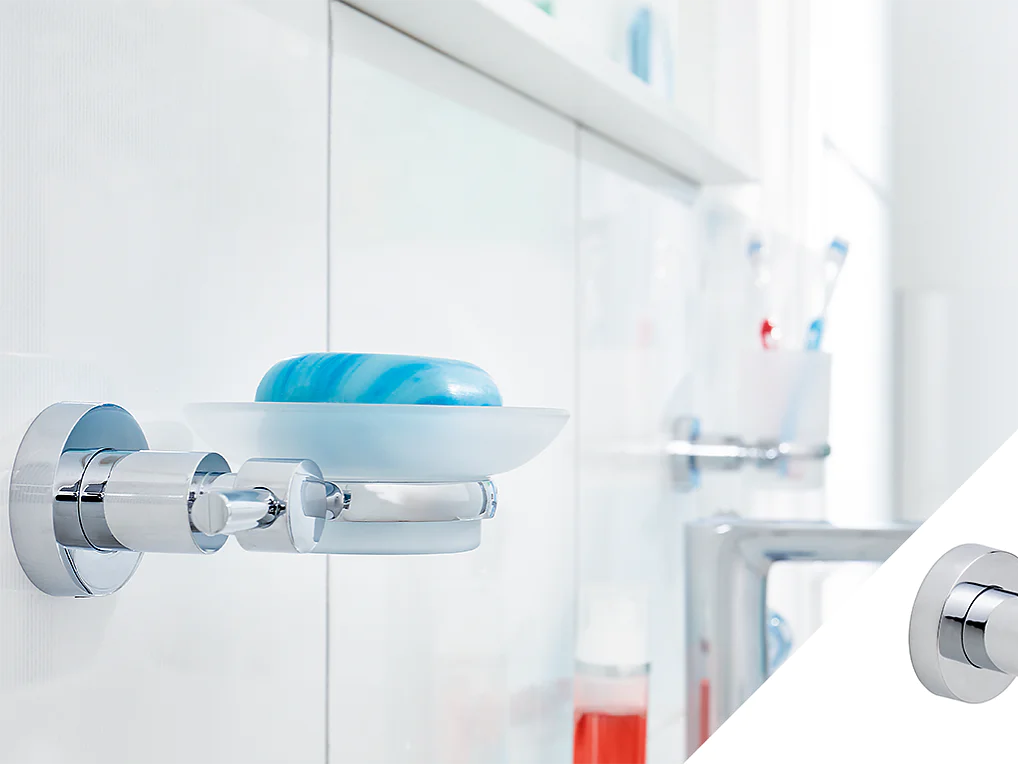 A chrome wall-mounted soap dish holds a blue bar of soap next to a white bathroom cabinet. In the corner, a separate chrome robe hook is displayed. Various toiletries are blurred in the background, and nearby, rolls of tesa tape are neatly stacked for convenient use. (This text has been generated by AI)