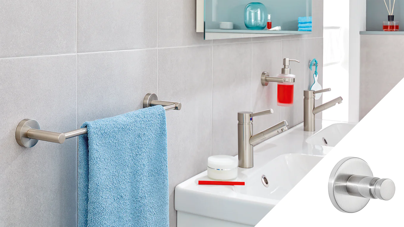 A modern bathroom features a double sink with two silver faucets and a red toothbrush. A blue towel hangs on a silver bar against a light gray tiled wall, secured by tesa tape. A small round silver wall hook is shown separately on the right, also attached using tesa tape. (This text has been generated by AI)