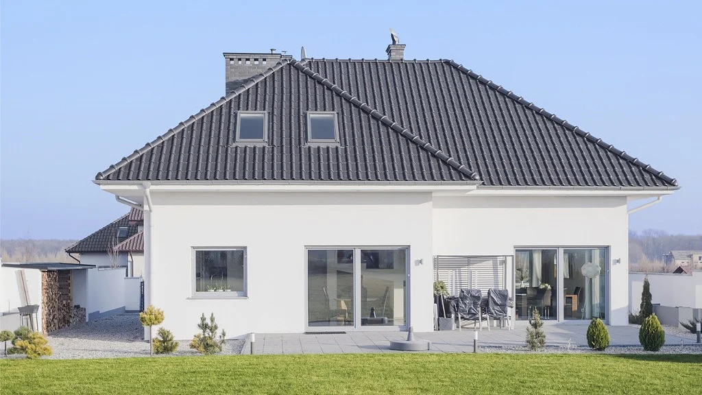Ein modernes weißes Haus mit einem dunkelgrauen Ziegeldach, zwei Dachfenstern und einem Schornstein. Das Haus hat große Fenster und einen Terrassenbereich mit Möbeln. Der Rasen vor dem Haus ist gut gepflegt und der Himmel ist klar. (This text has been generated by AI)
