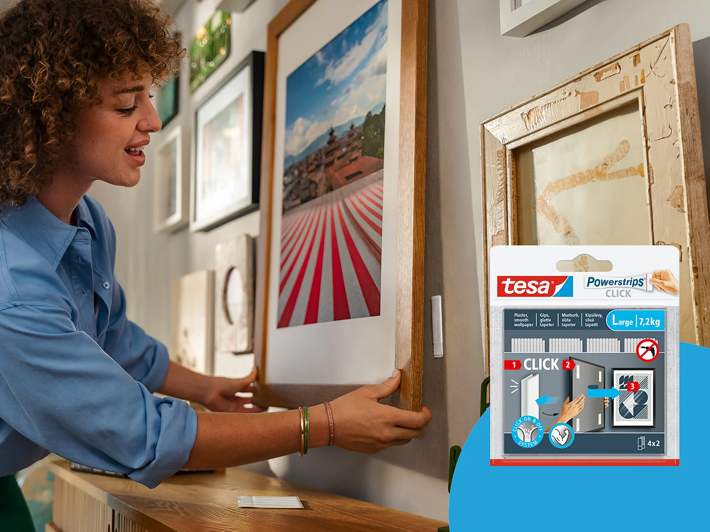A person with curly hair and a blue shirt hangs a framed artwork on a wall. Inset shows tesa tape Powerstrips packaging for hanging large items, indicating a weight capacity of 7.2 kg or 4 oz. Various other frames are displayed on the adjacent wall. (This text has been generated by AI)