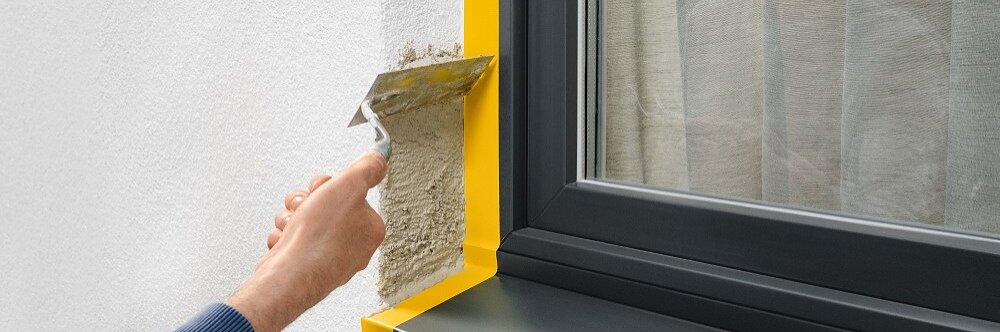 A person applies stucco with a trowel to a wall next to a window with a black frame and sill. The window has yellow tesa tape along the edges, and a reflection of the persons arm is visible in the glass. (This text has been generated by AI)