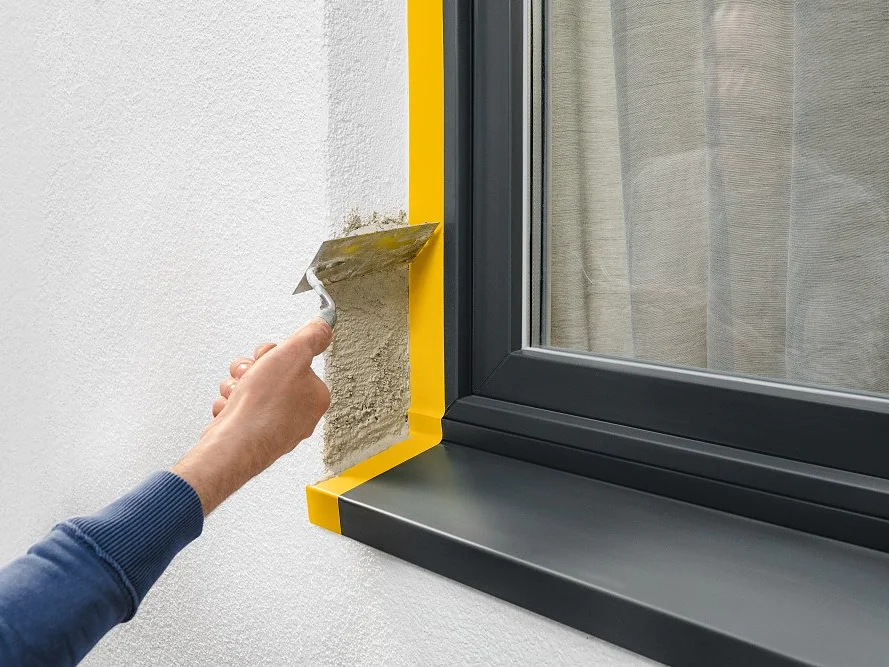 A person applies stucco with a trowel to a wall next to a window with a black frame and sill. The window has yellow tesa tape along the edges, and a reflection of the persons arm is visible in the glass. (This text has been generated by AI)