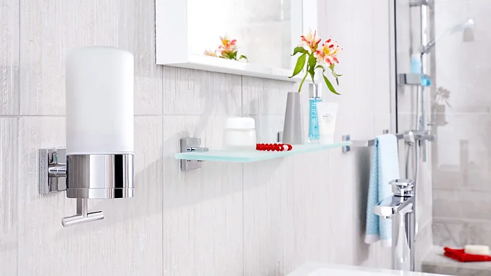 A modern bathroom features a wall-mounted soap dispenser and a glass shelf with toiletries, including a toothbrush, toothpaste, and a tube. A towel hangs on a rod secured with tesa tape, and a small bouquet of flowers is reflected in a wall-mounted mirror. (This text has been generated by AI)