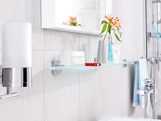A modern bathroom features a wall-mounted soap dispenser and a glass shelf with toiletries, including a toothbrush, toothpaste, and a tube. A towel hangs on a rod secured with tesa tape, and a small bouquet of flowers is reflected in a wall-mounted mirror. (This text has been generated by AI)