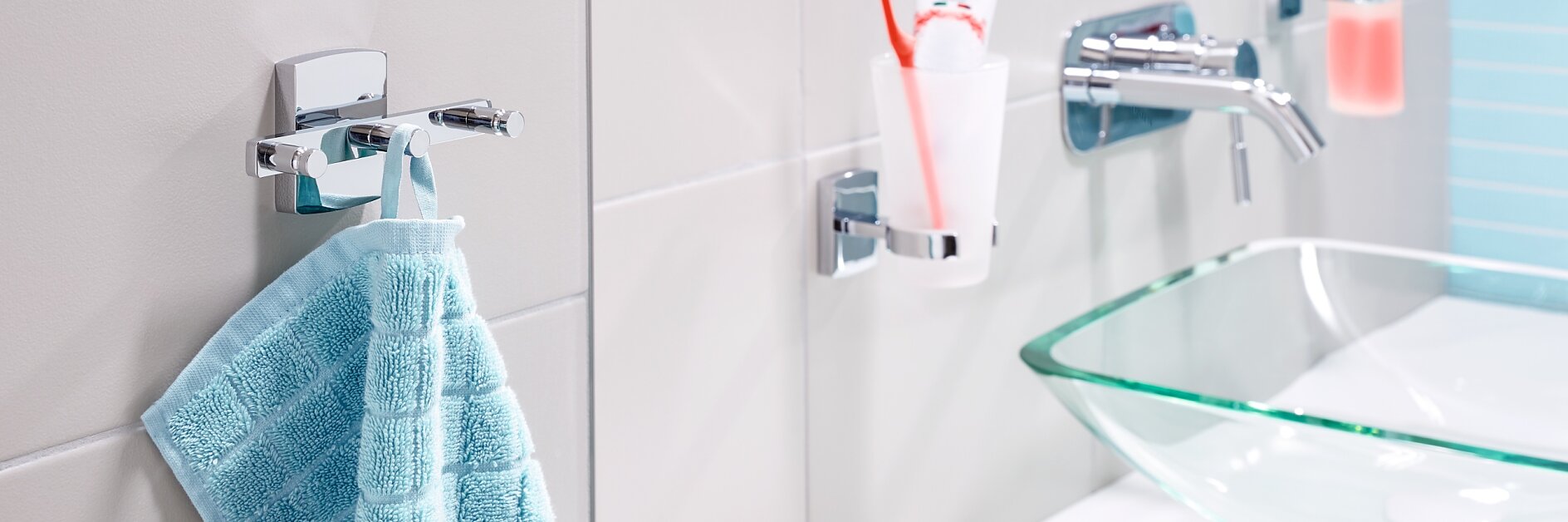 A modern bathroom features a light blue towel hanging on a wall hook near a clear glass sink with a chrome faucet. A toothbrush and toothpaste are in a holder next to a soap dispenser. The decor is minimalist with light-colored tiles. While the look is simple, clever use of tesa tape ensures secure placement of hooks and holders without compromising the clean aesthetic. (This text has been generated by AI)