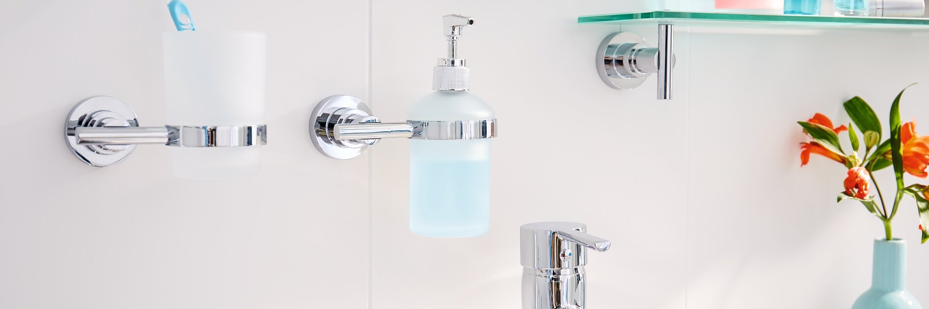 In the bathroom sink area, a chrome faucet complements a white counter. A toothbrush and holder, along with a soap dispenser, are securely mounted using tesa tape on the tiled wall. A shelf holding toiletries is also affixed with tesa tape. On the right side of the counter sits a small blue vase filled with orange flowers. (This text has been generated by AI)
