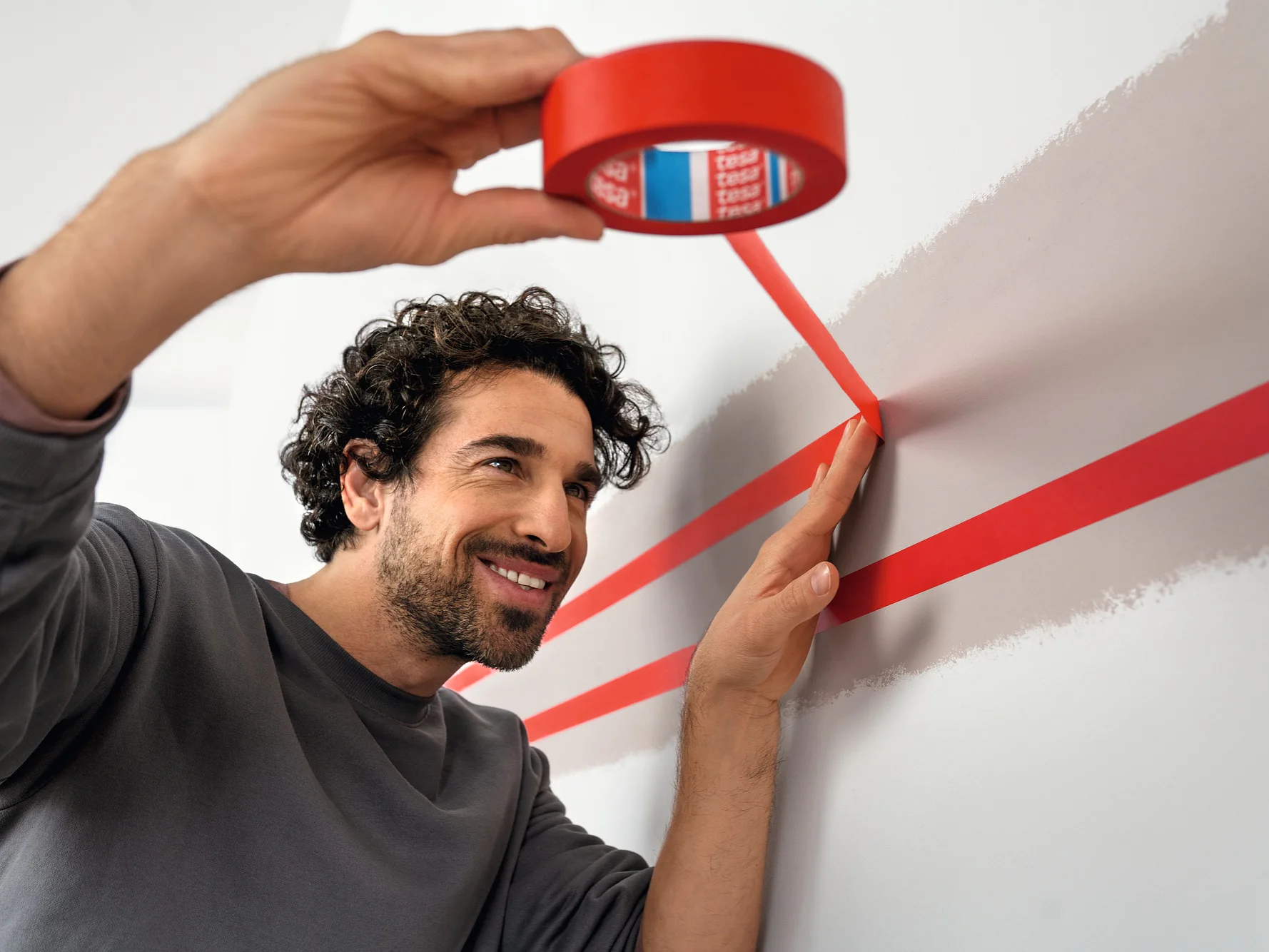 A person is applying red tesa tape to a wall, creating parallel lines. They are smiling and focused. The wall is light-colored, and the individual is wearing a gray long-sleeve shirt. The scene suggests preparation for painting or decoration. (This text has been generated by AI)