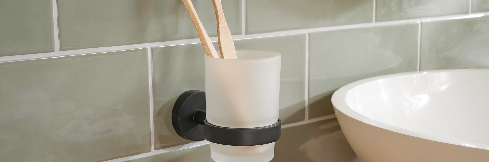 A bathroom scene featuring two wooden toothbrushes, one with white bristles and another with black bristles, in a frosted glass holder mounted on a light green tiled wall using tesa tape. A white sink is partially visible on a wooden countertop. (This text has been generated by AI)