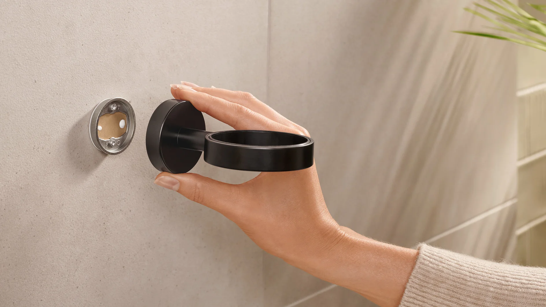 A hand is holding a black round bracket or holder against a wall. The wall has a circular mounting plate with screws visible, secured with tesa tape. The background is a light-toned tiled surface with part of a green plant in the top right corner. (This text has been generated by AI)