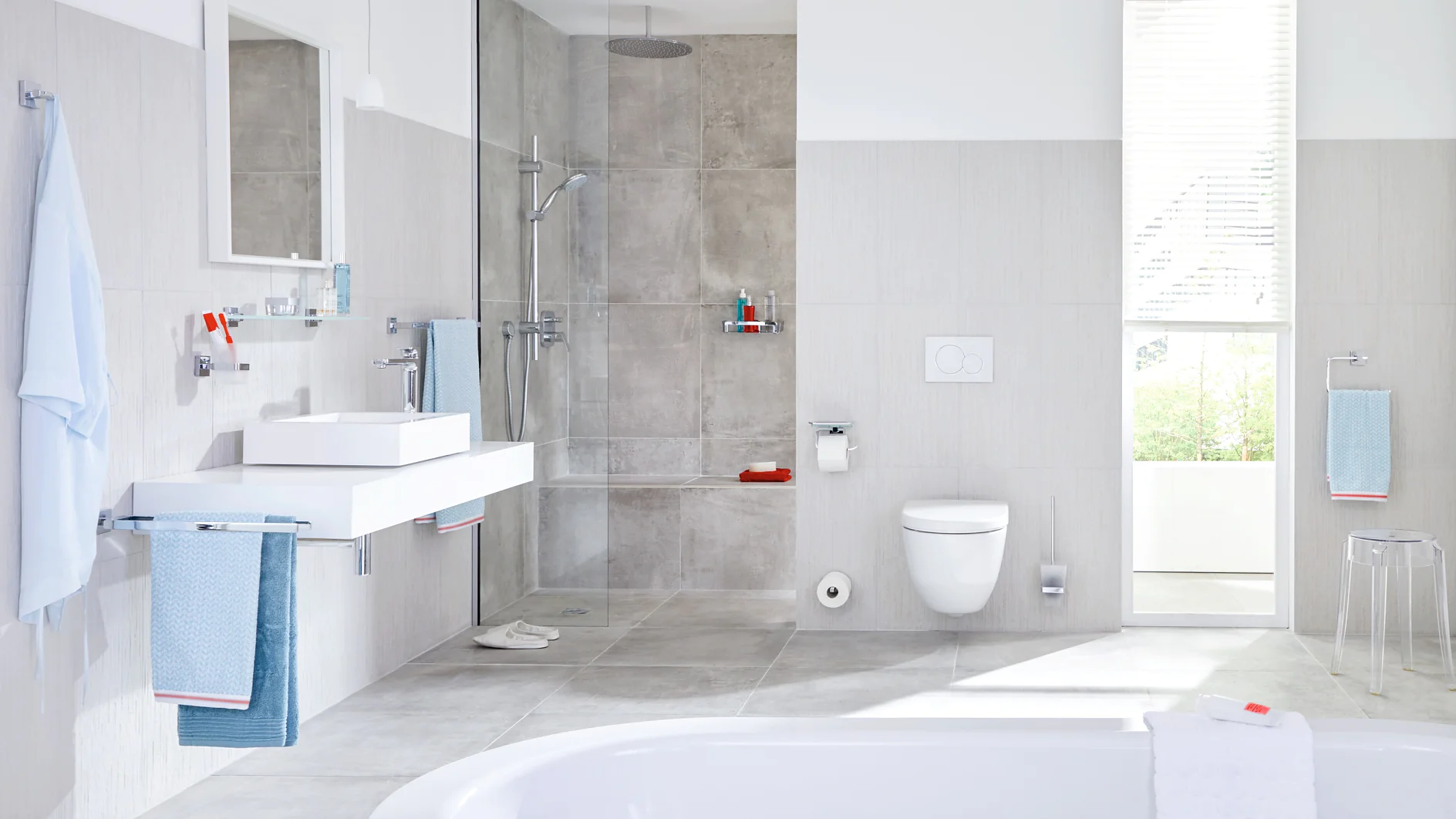Modern bathroom with a large tub in the foreground. On the left, a white sink with a mirror above. The shower area has gray tiles and a glass door. A white toilet is next to the shower. A large window provides natural light, and towels hang nearby using tesa tape. (This text has been generated by AI)