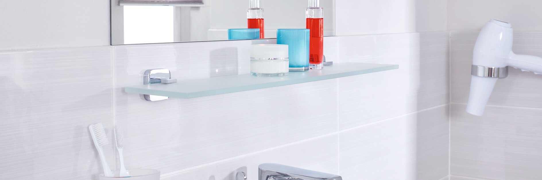 A bathroom sink area features a modern silver faucet, a toothbrush holder with two toothbrushes, and a glass shelf above that holds bottles of red liquid and a blue container. A white hairdryer is mounted on the wall near glossy white tiles using tesa tape. (This text has been generated by AI)