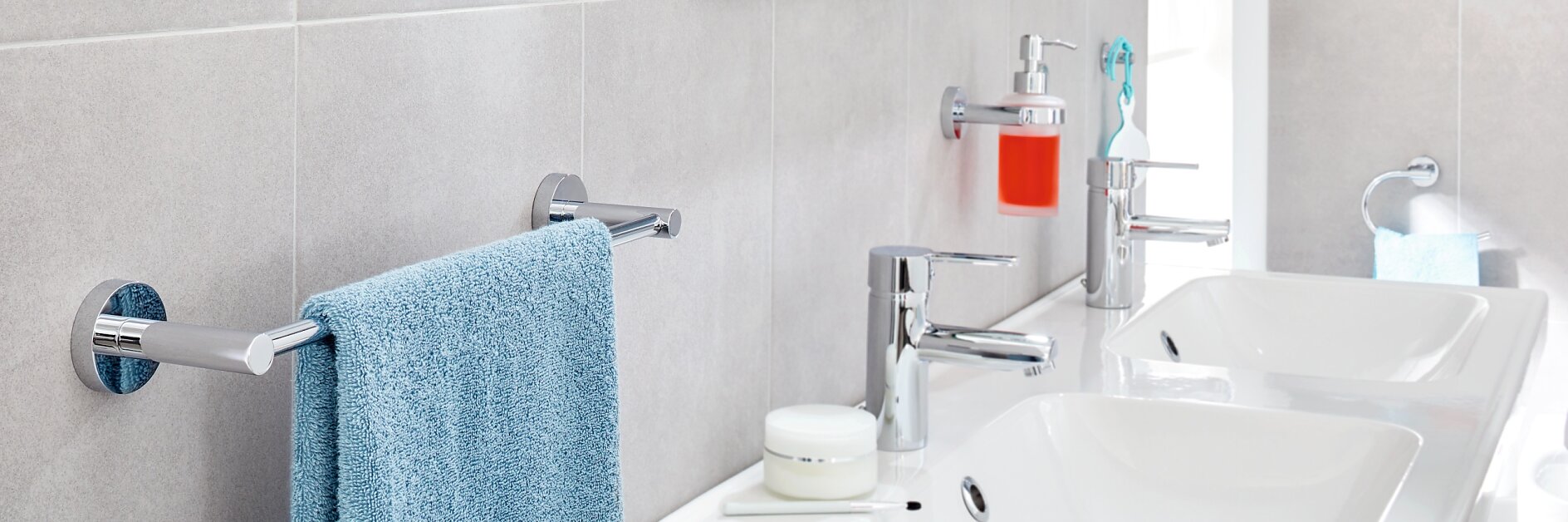 A modern bathroom features two white sinks with chrome faucets on a countertop. A blue towel hangs on a nearby towel bar, secured with tesa tape. A bright red soap dispenser is mounted on the wall using tesa tape. Shelves above are stocked with toiletries and towels, neatly held in place by tesa tape. (This text has been generated by AI)