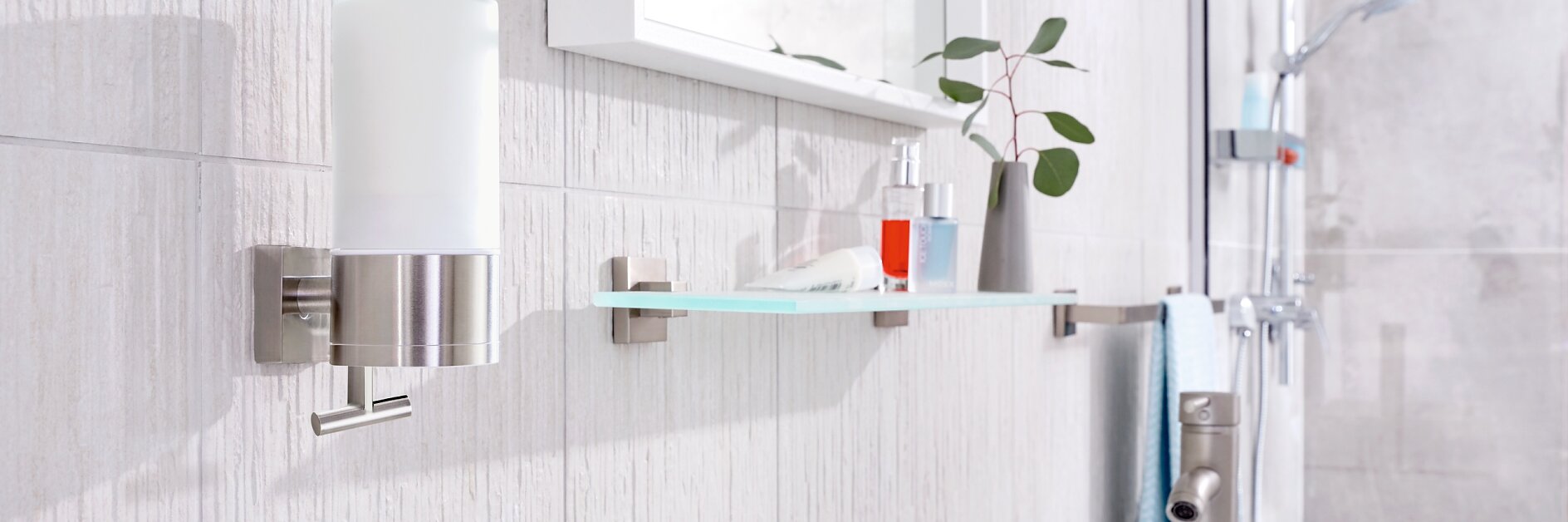 A modern bathroom with a wall-mounted glass shelf holding a small plant and red bottle, all secured using tesa tape. A soap dispenser is mounted nearby on a tiled wall. The sink faucet is visible below, and a shower head is seen in the background. (This text has been generated by AI)