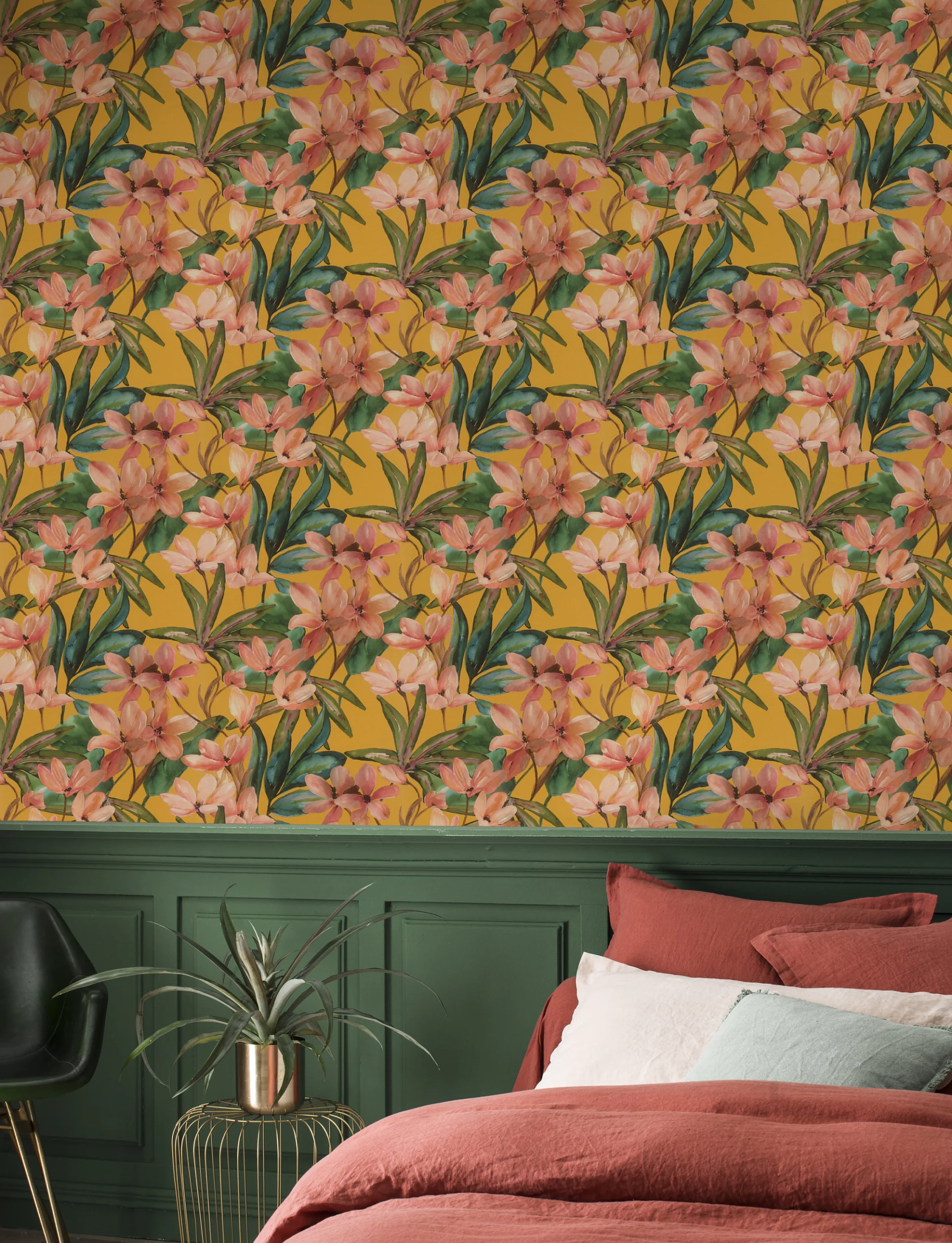 A bedroom with a bed featuring rust-colored bedding, a green wainscoted wall, and wallpaper patterned with large pink flowers and green leaves on a yellow background, installed using tesa tape. A black chair and a potted plant are also visible. (This text has been generated by AI)