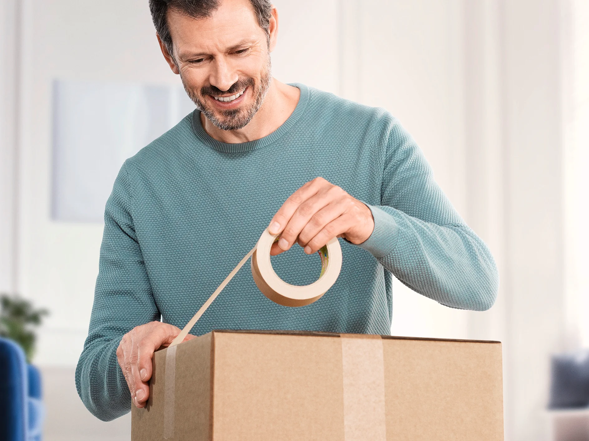 A man in a teal sweater is sealing a cardboard box with tesa tape. He is smiling and appears to be indoors with a blurred background. (This text has been generated by AI)