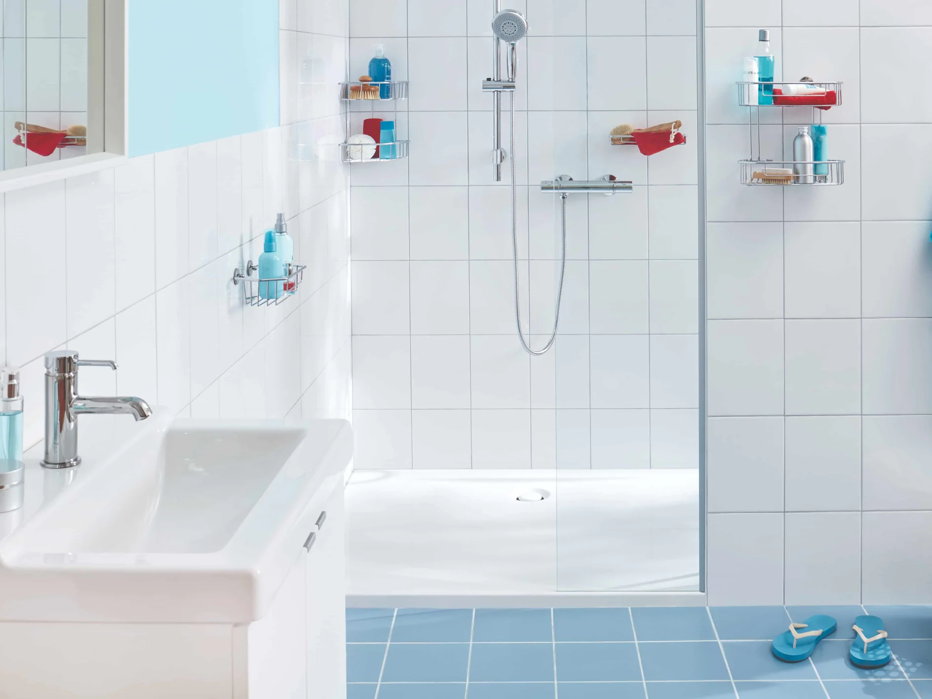 A modern bathroom features white tile walls and a white sink. The shower area includes a glass divider, a handheld showerhead, and shelves for toiletries. A blue towel is neatly hung on the right, accompanied by matching slippers and a laundry basket on the blue-tiled floor. (This text has been generated by AI)