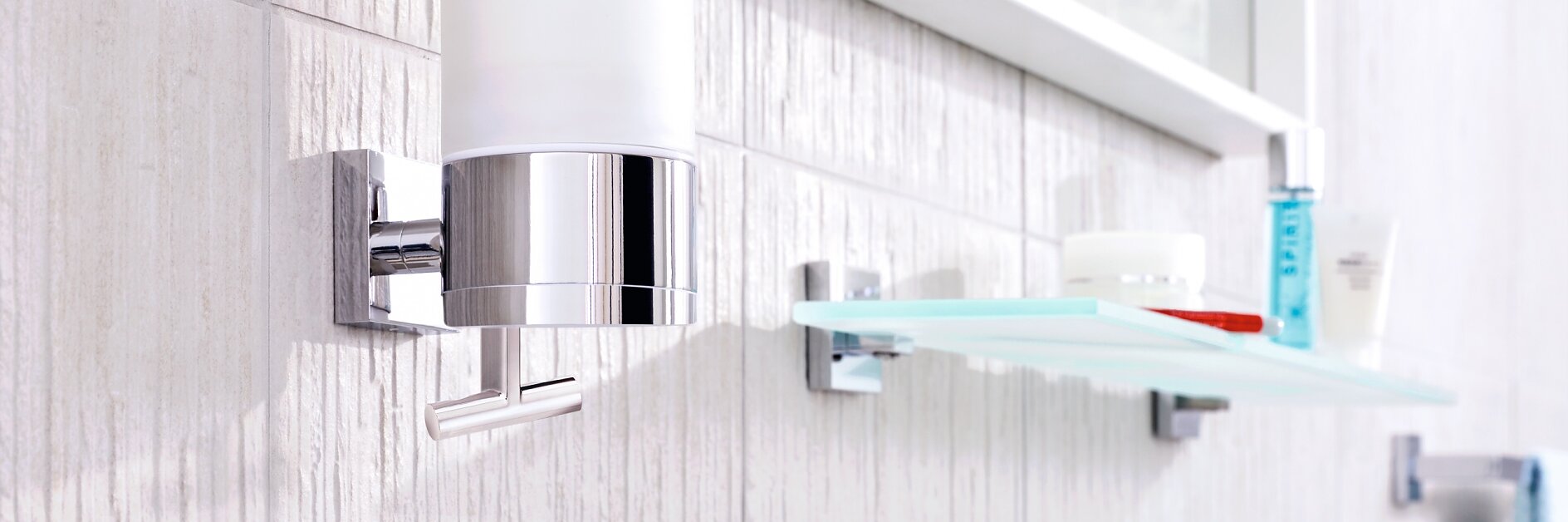 A bathroom with a modern soap dispenser mounted on a textured white wall using tesa tape. Below it is a glass shelf secured with tesa tape, holding various toiletry items. The reflection of the shelf can be seen in a nearby mirror. (This text has been generated by AI)