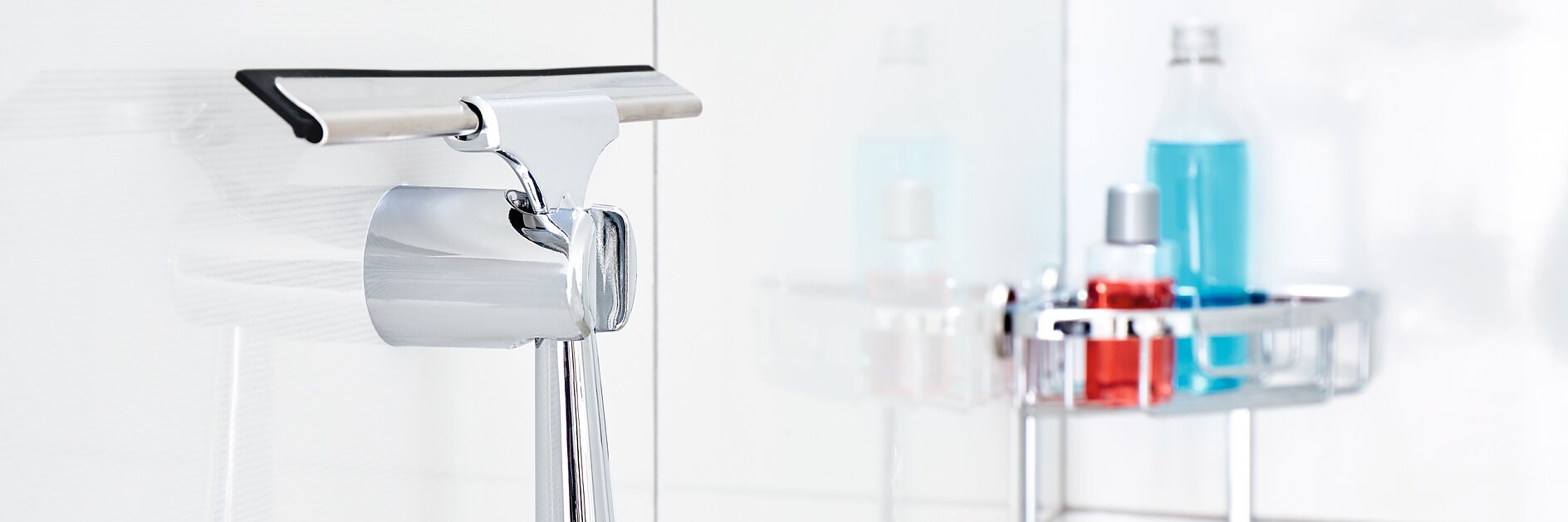 A chrome squeegee hangs on a white tiled bathroom wall, fastened securely with tesa tape. In the background, there are shower caddies holding a blue bottle, a red bottle, and a yellow sponge. (This text has been generated by AI)