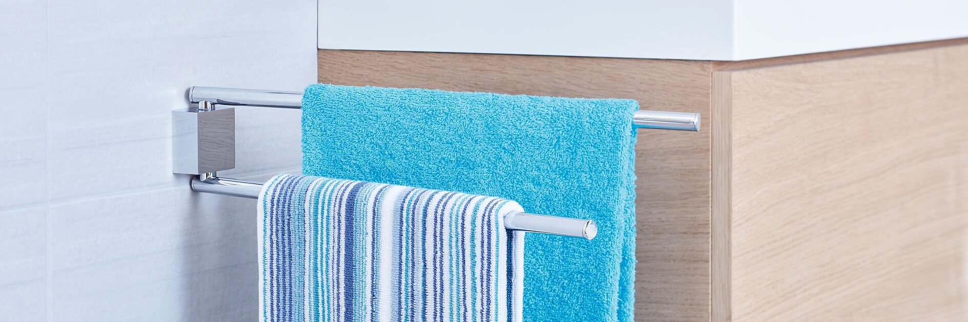 A modern bathroom features a white sink with a chrome faucet, set on a wooden cabinet. In the foreground, a towel rack holds a blue towel and a striped towel, mounted on the tile wall using tesa tape. (This text has been generated by AI)