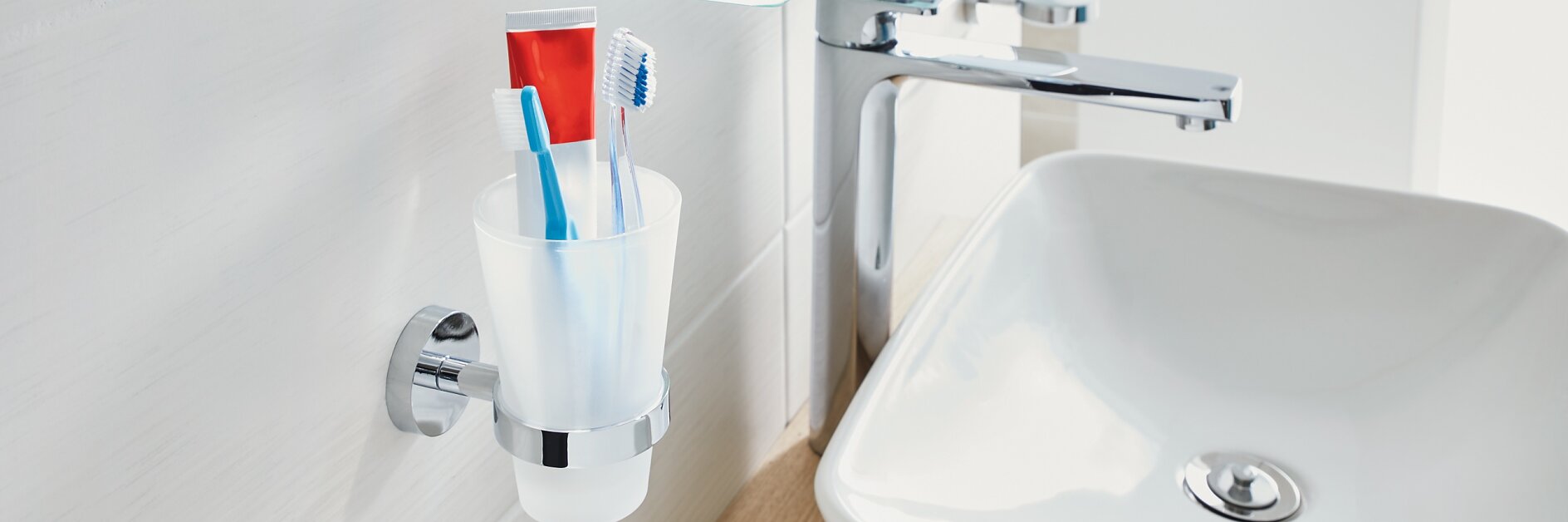 A bathroom sink area featuring a white basin with a silver faucet. A glass shelf above holds toiletries like cologne and creams. A wall-mounted cup contains two toothbrushes and a tube of toothpaste secured neatly in place using tesa tape. The background is white and clean. (This text has been generated by AI)
