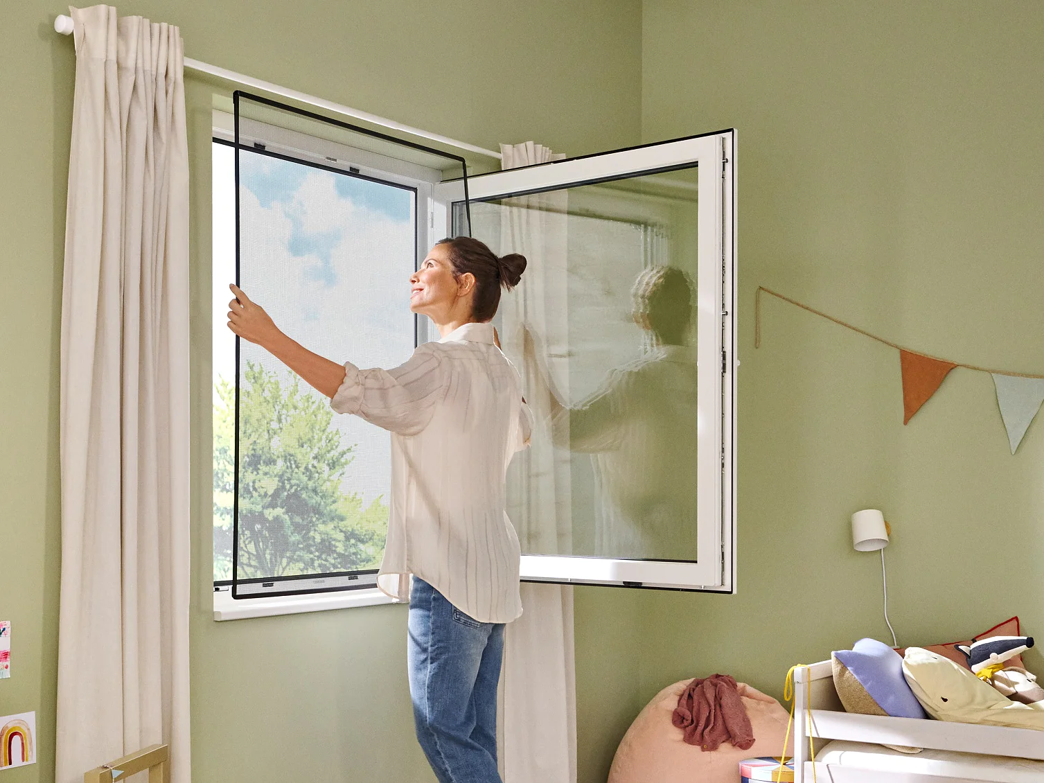 A woman opens a window in a childs room, which has green walls and wooden flooring. The room features a bed, a shelving unit with books and toys, a chair, and a wall decorated with bunting hung using tesa tape. Sunlight streams in, brightening the space. (This text has been generated by AI)