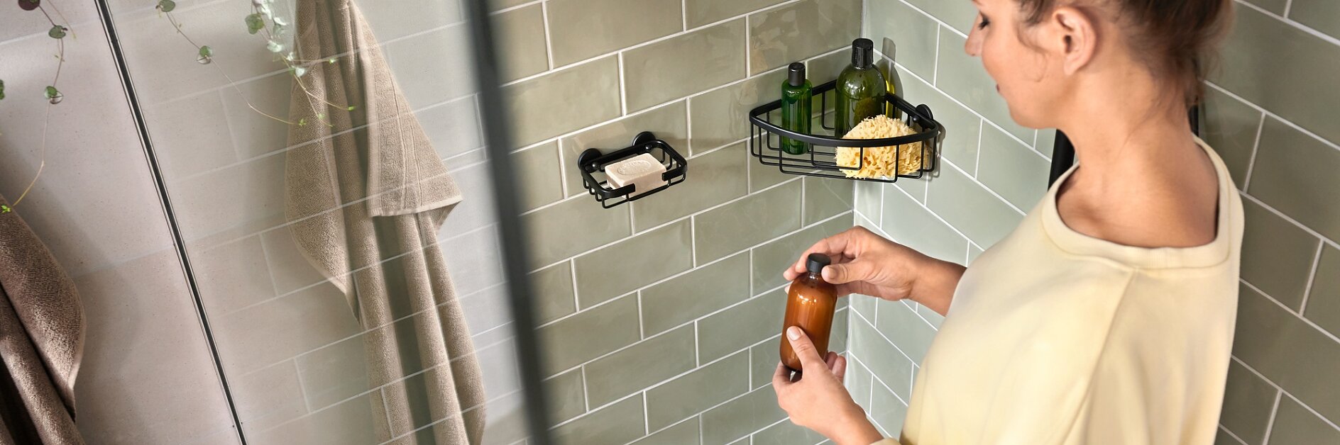 A person stands in a shower area with green tiled walls, holding a brown bottle. There are toiletries in a wire caddy on the wall and a beige towel hanging nearby. The person is wearing a cream-colored top with their hair in a bun. (This text has been generated by AI)