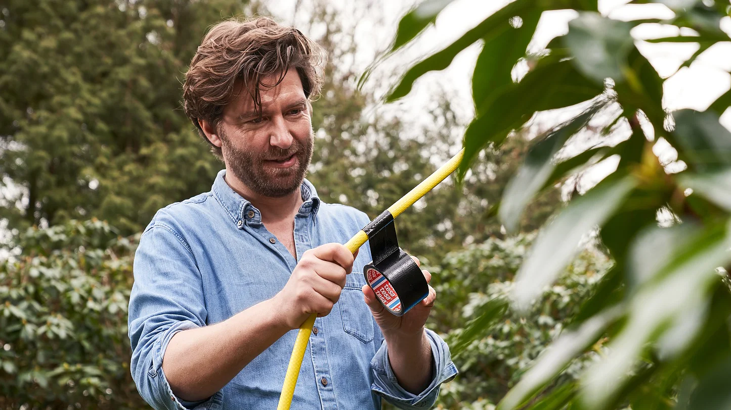 A bearded man in a blue shirt is standing outdoors, holding a yellow hose and a roll of tesa tape. He appears to be repairing or adjusting the hose. Green foliage and trees surround him in the background. (This text has been generated by AI)
