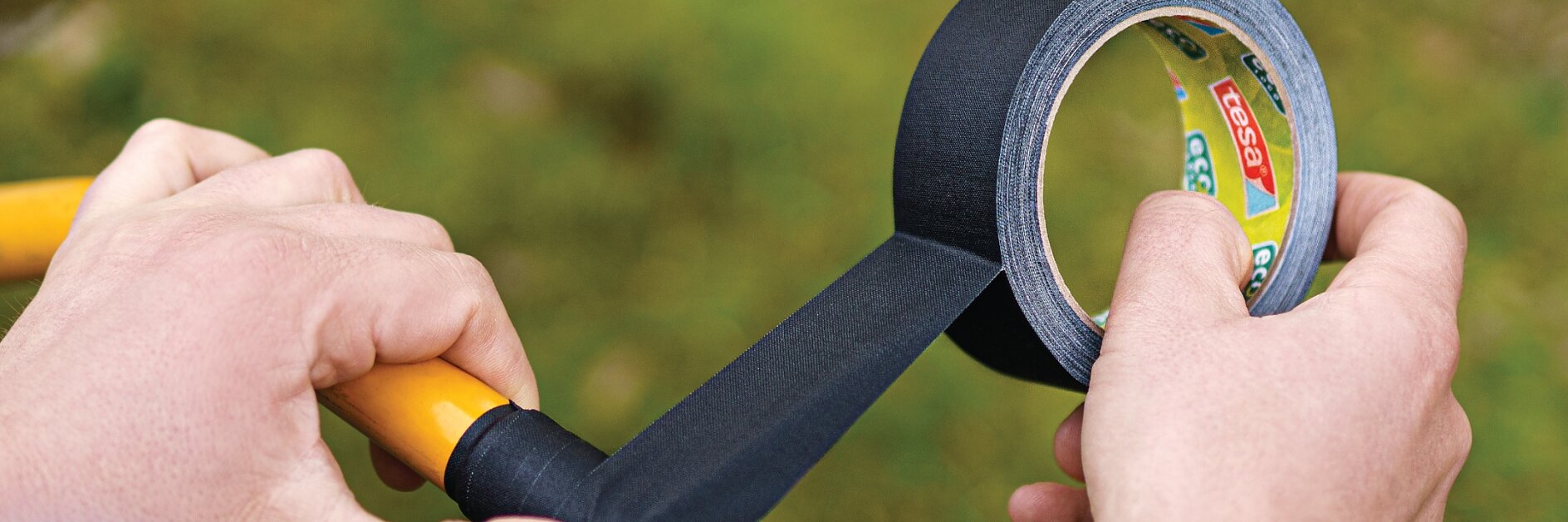 A person wraps black tesa tape around the handle of a yellow tool. The tesa tape is being unrolled from a branded roll. The background is blurred with green and brown tones, suggesting an outdoor setting. (This text has been generated by AI)