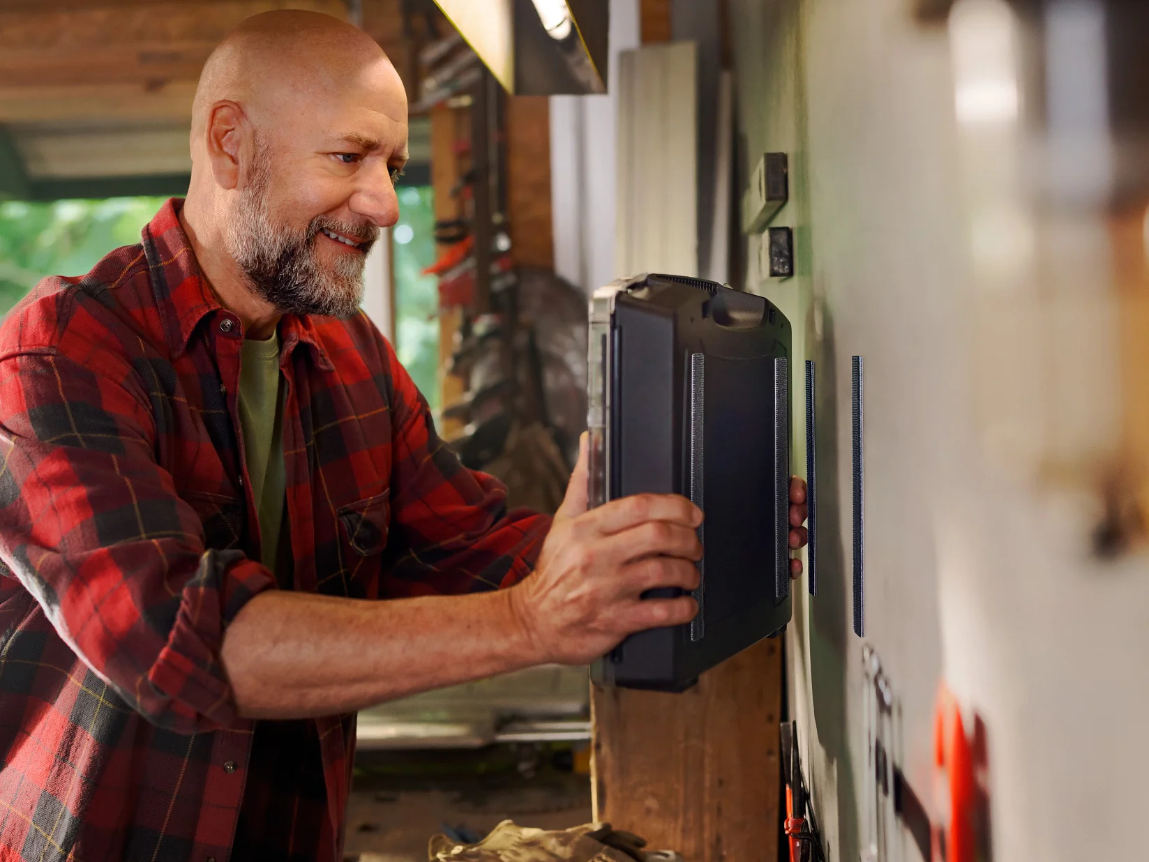 A bald man with a beard, wearing a red plaid shirt, is placing a toolbox on a shelf in his workshop. He smiles as he organizes tools around him. The workshop is brightly lit with various tools and equipment visible in the background, along with tesa tape prominently displayed among them. (This text has been generated by AI)