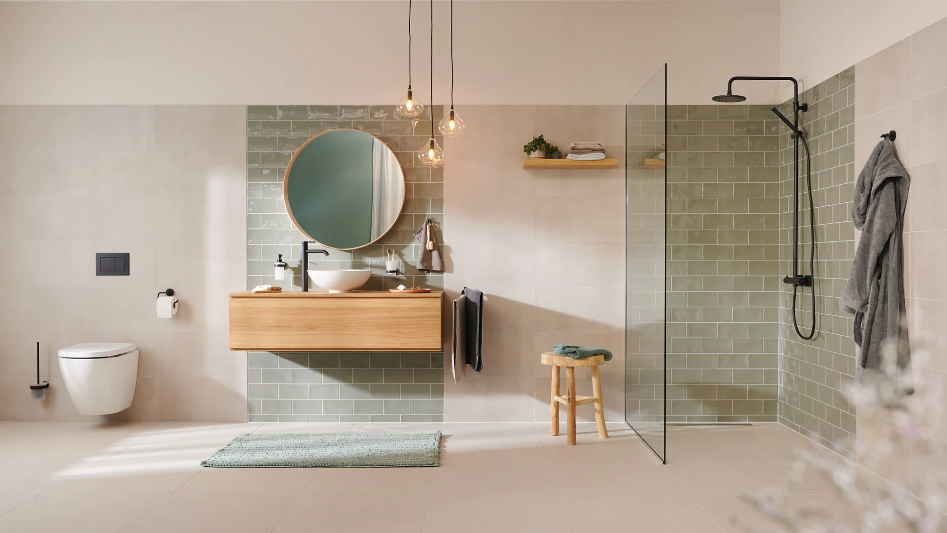 A modern bathroom with beige tiles and a green tiled accent wall. Features include a round mirror above a wooden vanity with a white sink, a walk-in shower with black fixtures, a wall-mounted toilet, pendant lights, and a towel on a hook secured with tesa tape. (This text has been generated by AI)