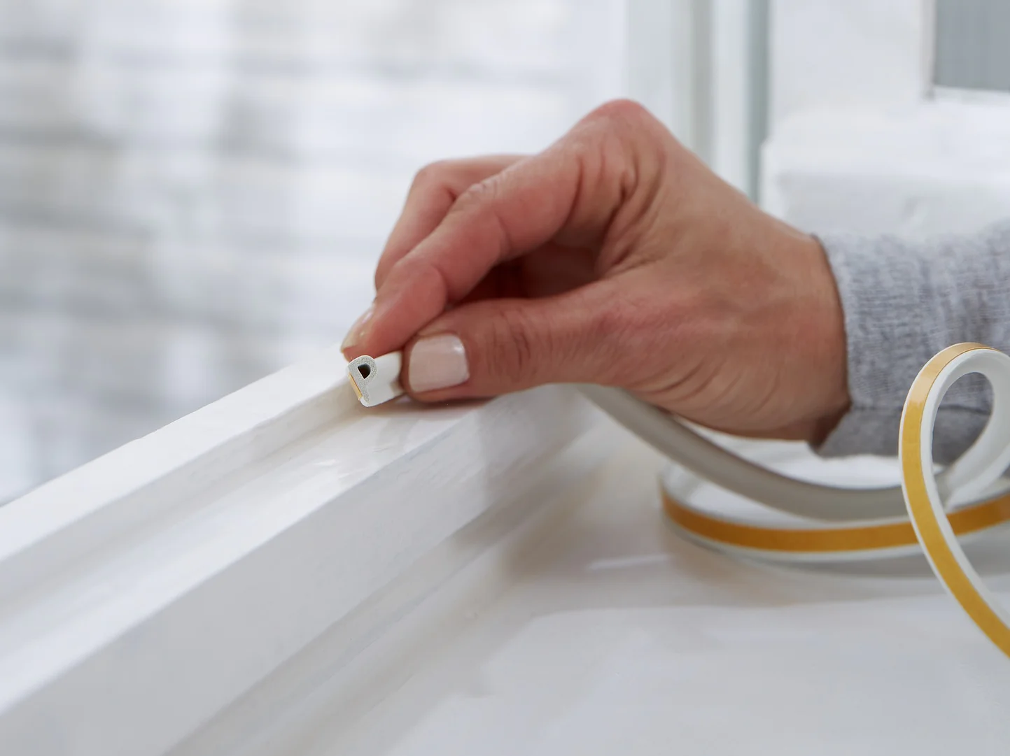 A hand is placing tesa tape weather stripping onto a white window frame. The background shows a blurred exterior wall. The weather stripping is beige with a peel-off backing, suggesting a home improvement or insulation task. (This text has been generated by AI)