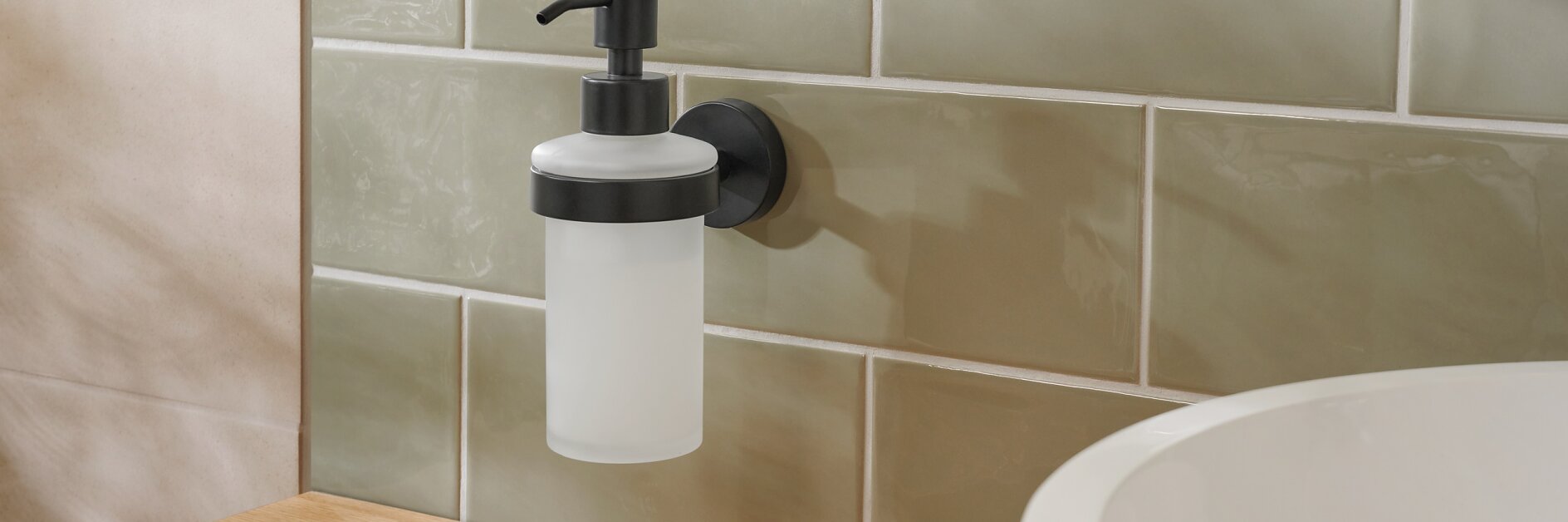 Ein wandmontierter Seifenspender mit schwarzer Pumpe ist mit Tesa-Klebeband an einer gefliesten Wand befestigt. Die Fliesen haben einen gedeckten Grünton. Der Spender befindet sich über einer Holzarbeitsplatte mit einer rosa Schale und neben einem weißen Keramikwaschbecken. (This text has been generated by AI)