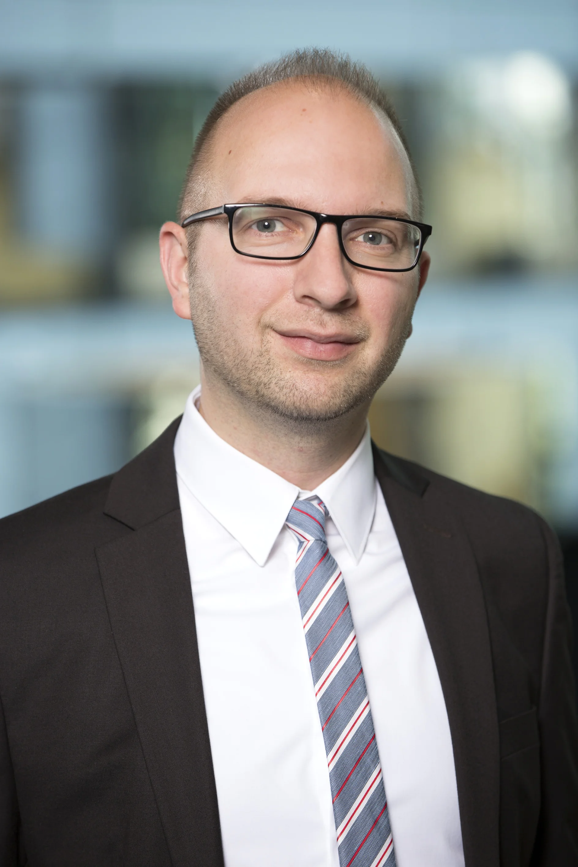 Eine Person mit kurzen Haaren, Brille, schwarzem Anzug, weißem Hemd und gestreifter Krawatte steht vor einem unscharfen Innenhintergrund. (This text has been generated by AI)