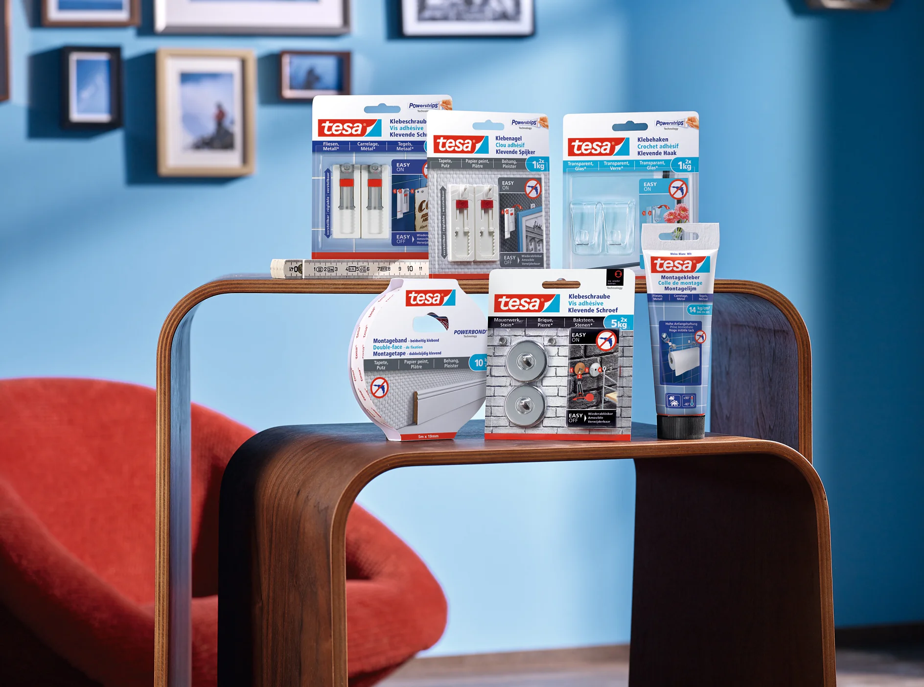 tesa_Cleveres_Befestigungs-System_Produkt-Highlights.tif Auf einem Holzregal vor einer blauen Wand sind verschiedene Klebeprodukte von tesa ausgestellt. Zu den Artikeln gehören Klebestreifen, Haken, Montagebänder und Schutzpolster. Im Hintergrund sind ein roter Stuhl und gerahmte Bilder zu sehen. (This text has been generated by AI)
