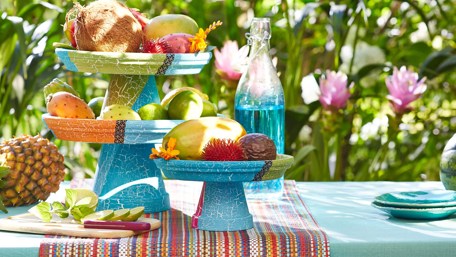 Auf einem farbenfrohen Tisch im Freien stehen Obstsorten in zwei blauen Schalen. Zu den Früchten gehören Kokosnüsse, Mangos und Zitrusfrüchte. Daneben stehen eine Ananas, eine Wassermelone und eine blaue Glasflasche. Im Hintergrund sind grüne Blätter und rosa Blüten zu sehen. (This text has been generated by AI)