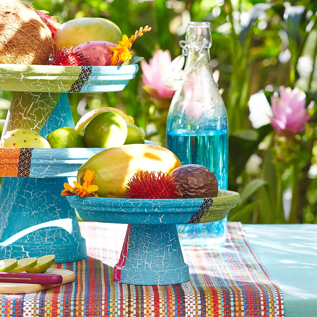Auf einem farbenfrohen Tisch im Freien stehen Obstsorten in zwei blauen Schalen. Zu den Früchten gehören Kokosnüsse, Mangos und Zitrusfrüchte. Daneben stehen eine Ananas, eine Wassermelone und eine blaue Glasflasche. Im Hintergrund sind grüne Blätter und rosa Blüten zu sehen. (This text has been generated by AI)