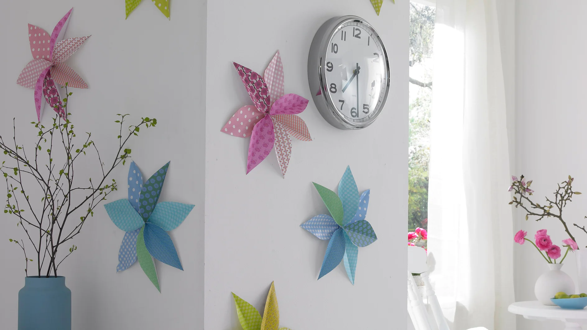 An einer weißen Wand hängt eine Wanduhr, die mit Windrädern aus Papier in rosa, blauen, grünen und gelben Mustern dekoriert ist. Links steht eine blaue Vase mit Zweigen und rechts ein Fenster mit transparenten Vorhängen, das natürliches Licht hereinlässt. (This text has been generated by AI)