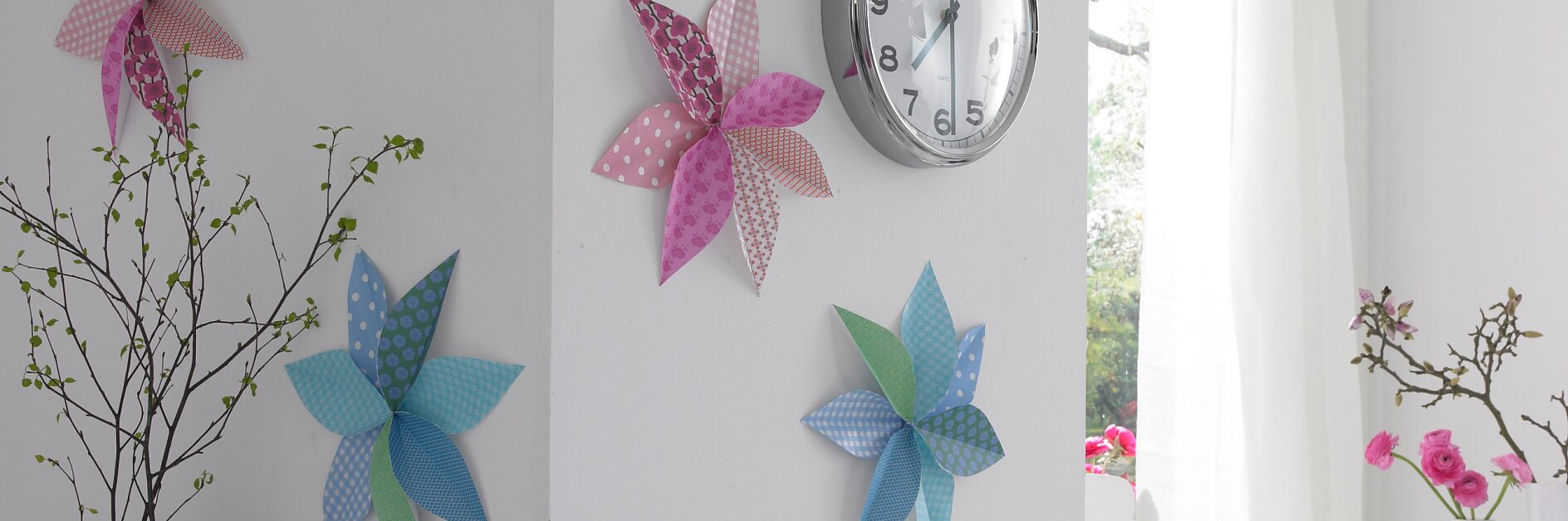 An einer weißen Wand hängt eine Wanduhr, die mit Windrädern aus Papier in rosa, blauen, grünen und gelben Mustern dekoriert ist. Links steht eine blaue Vase mit Zweigen und rechts ein Fenster mit transparenten Vorhängen, das natürliches Licht hereinlässt. (This text has been generated by AI)