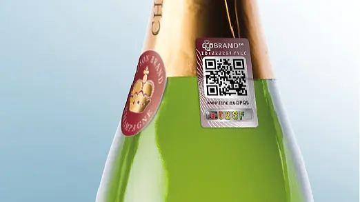 Nahaufnahme einer Champagnerflasche mit einer grünlichen Flüssigkeit im Inneren. Der Flaschenhals zeigt ein Etikett mit einem Kronenlogo und einem metallischen QR-Code-Aufkleber mit alphanumerischen Zeichen und einer Webadresse, der mit Tesa-Klebeband versiegelt ist. Der Hintergrund ist ein hellblauer Farbverlauf.