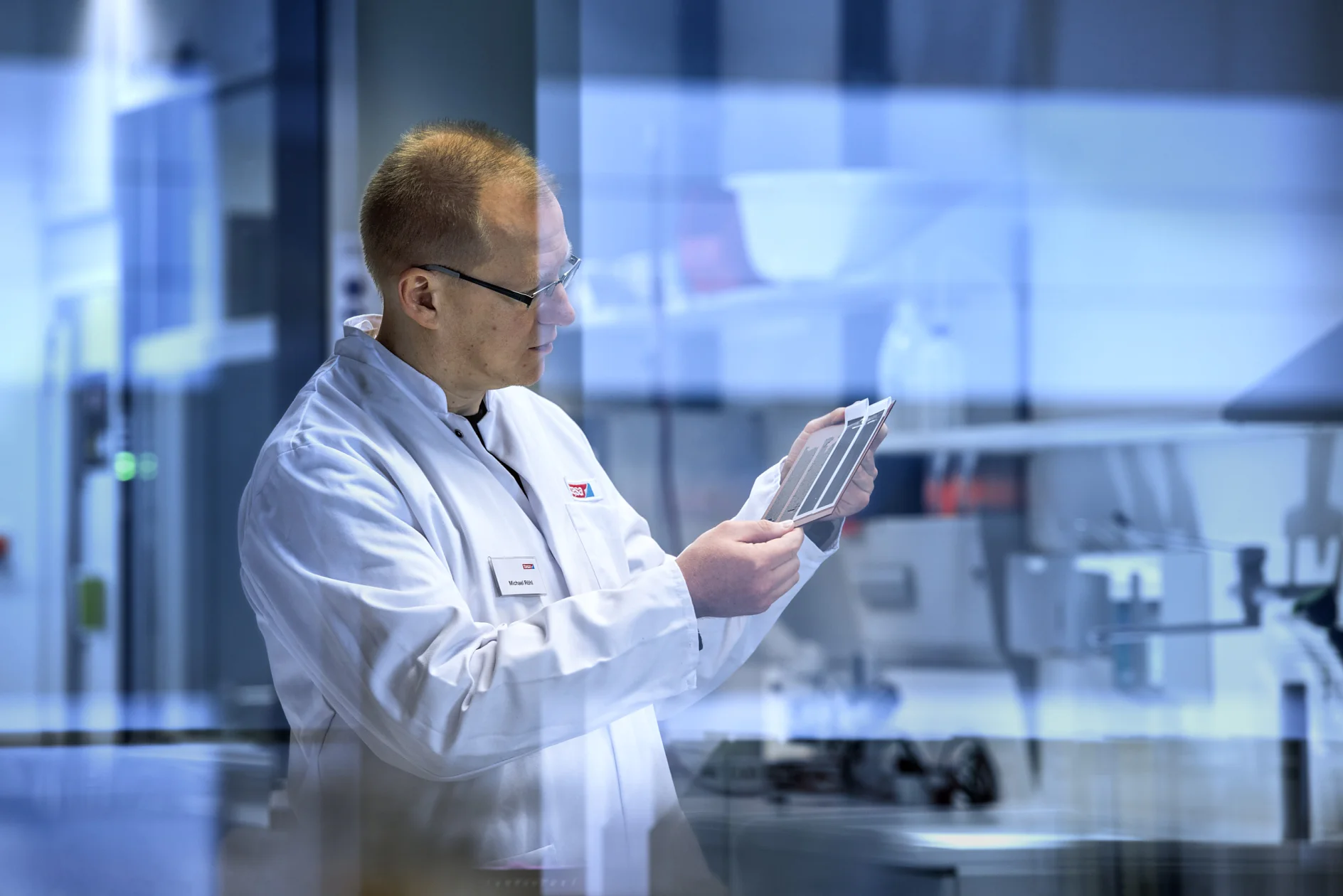 tesa_Forscher.jpg Eine Person im Laborkittel untersucht in einer Laborumgebung ein Blatt mit Testproben. Die Umgebung ist modern mit Glas- und Metallelementen, und im Hintergrund sind verschiedene Laborgeräte, darunter Tesa-Klebeband, zu sehen. (This text has been generated by AI)