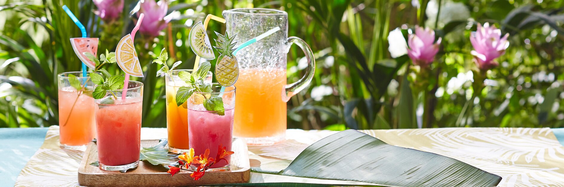 Eine tropische Außenkulisse mit drei bunten Fruchtcocktails und einem Krug Saft auf einem Tisch. Jedes Getränk ist mit Fruchtscheiben und Minze garniert. Der Tisch ist mit einem Bananenblatt und Blumen dekoriert. Im Hintergrund sind üppiges Grün und Blumen zu sehen. (This text has been generated by AI)