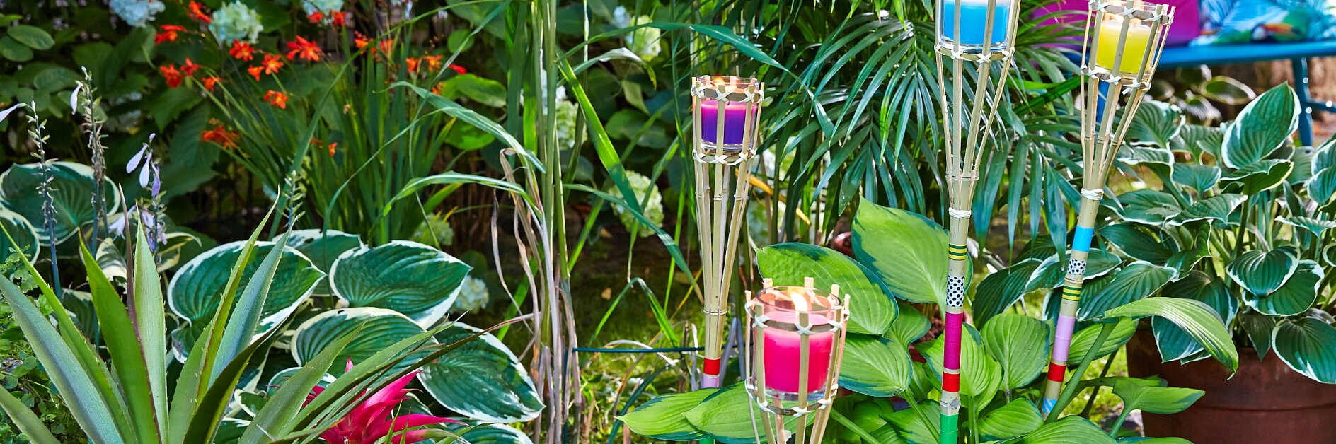 Eine Gartenszene mit üppigem grünem Laub und Pflanzen. Bunte Tiki-Fackeln mit rosa, violetten, blauen und gelben Spitzen sind zwischen den Pflanzen platziert. Im Hintergrund sind auch Blumen in den Farben Rot, Rosa und Weiß zu sehen. (This text has been generated by AI)
