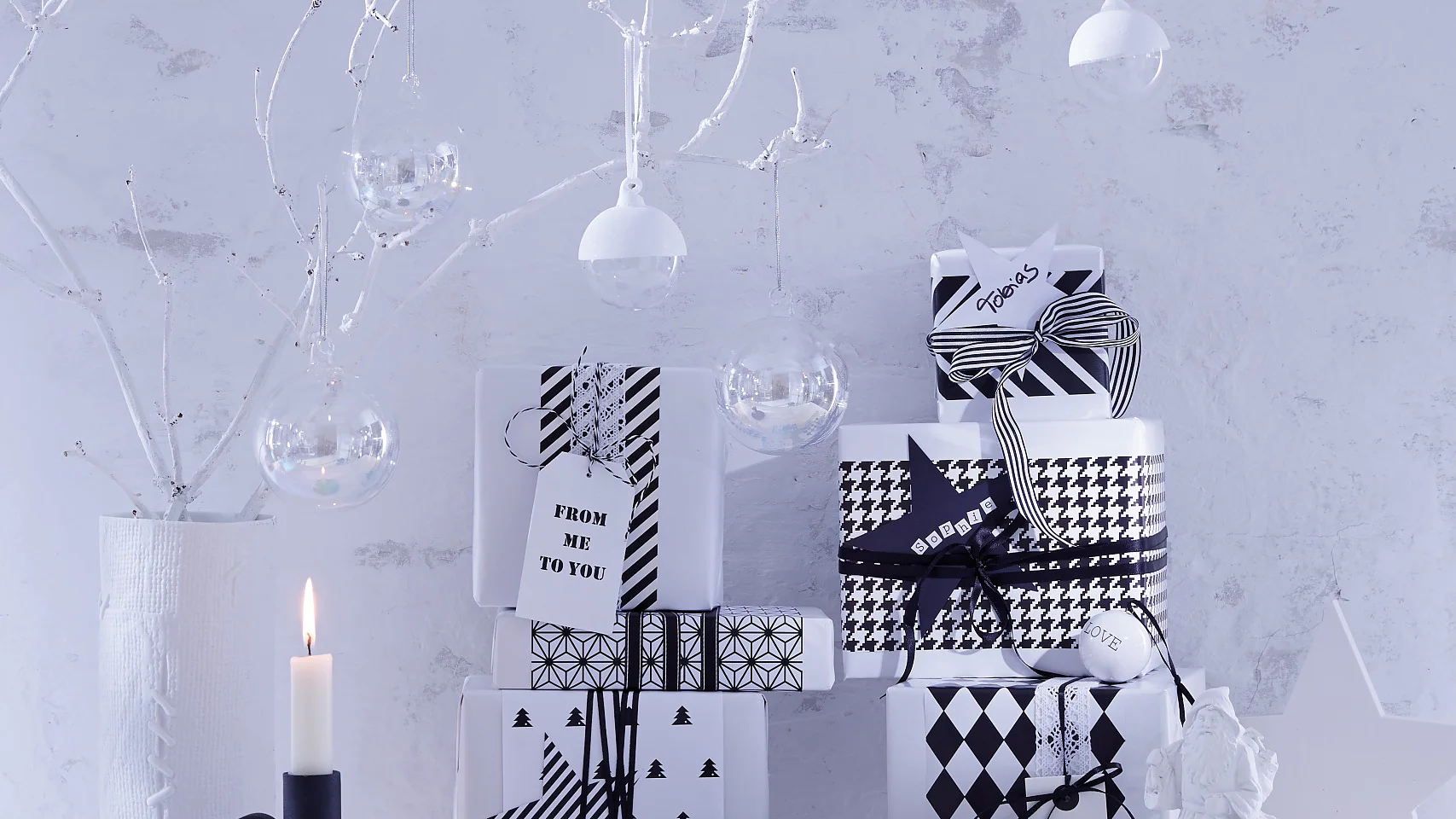 Eine minimalistische Winterlandschaft mit schwarz-weiß verpackten Geschenken, die mit Tesa-Klebeband auf einer weißen Oberfläche gestapelt sind. Daneben steht ein beleuchteter schwarzer Kerzenständer. Weiße Zweige mit hängenden Ornamenten und kleine leuchtende Kerzen schaffen vor einem blassen Hintergrund eine heitere, monochrome Atmosphäre.