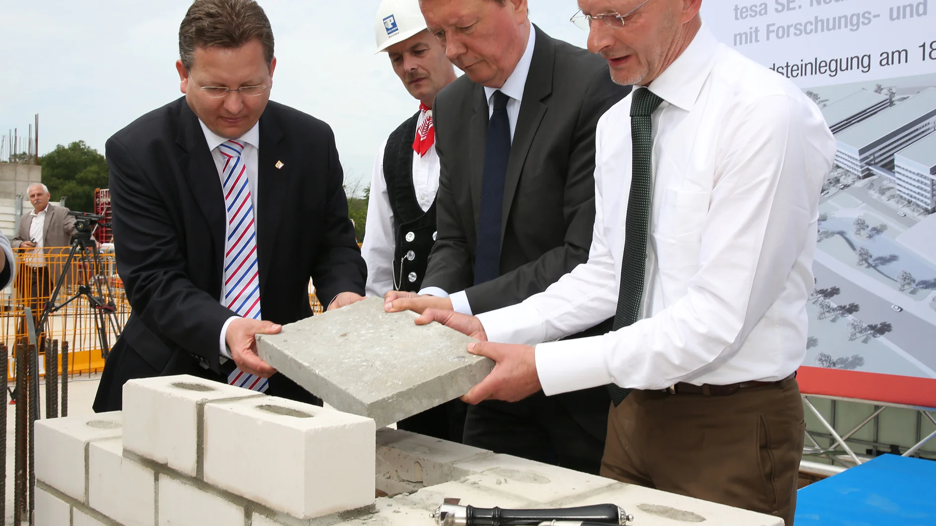 Vier Männer in formeller Kleidung nehmen an einer Maurerzeremonie teil. Einer trägt einen Bauhelm, während zwei andere eine Betonplatte über eine teilweise errichtete Mauer halten. Im Hintergrund sind eine Maurerkelle und ein Schild zu sehen, und mehrere Rollen Tesa-Klebeband sind strategisch auf der Baustelle verteilt.
