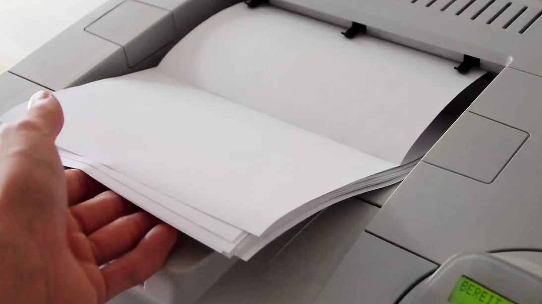 Eine Person hält einen Stapel weißes Papier in der Hand, der in einen grauen Drucker eingelegt werden soll. Auf dem Display des Druckers leuchtet ein grünes Licht und das Wort „Bereit“, was bedeutet, dass der Drucker bereit ist. Tesa-Klebeband.