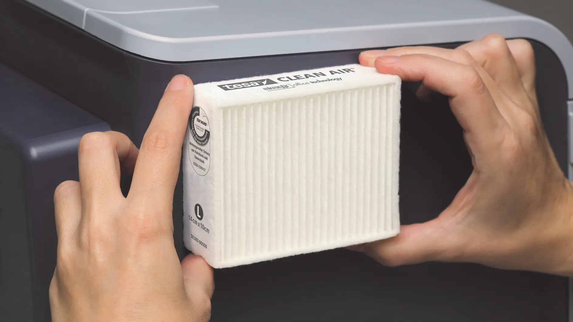 Hände setzen einen weißen rechteckigen Luftfilter mit der Aufschrift „CLEAN AIR“ in ein Gerät ein. Der Filter hat sichtbare Rillen und ist mit Text beschriftet. Das Gerät ist überwiegend dunkel mit einer hellgrauen Oberseite, und die Kanten des Luftfilters sind mit Tesa-Klebeband befestigt.