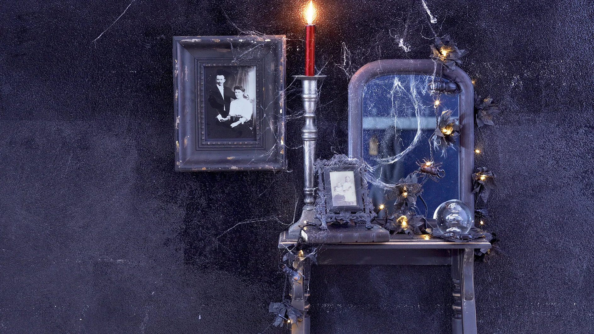Ein kleiner Vintage-Tisch mit einem gerahmten Schwarzweißfoto, einer brennenden roten Kerze und einem mit Spinnweben bedeckten Spiegel. Die Szene ist mit einer Reihe schwacher Lichterketten und künstlichen Spinnen dekoriert, die alle kreativ mit Tesa-Klebeband an einer dunklen, strukturierten Wand aufgehängt sind. (This text has been generated by AI)