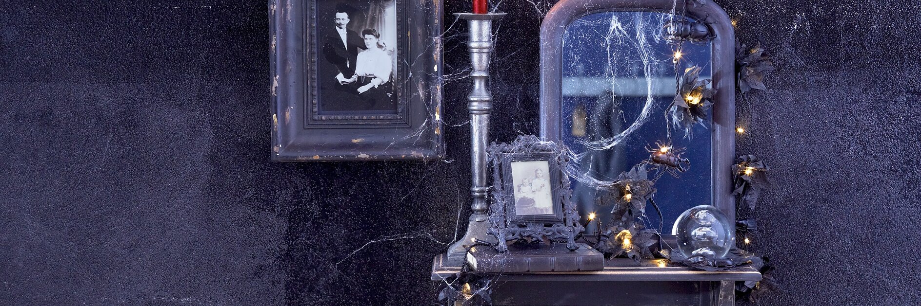 Ein kleiner Vintage-Tisch mit einem gerahmten Schwarzweißfoto, einer brennenden roten Kerze und einem mit Spinnweben bedeckten Spiegel. Die Szene ist mit einer Reihe schwacher Lichterketten und künstlichen Spinnen dekoriert, die alle kreativ mit Tesa-Klebeband an einer dunklen, strukturierten Wand aufgehängt sind. (This text has been generated by AI)
