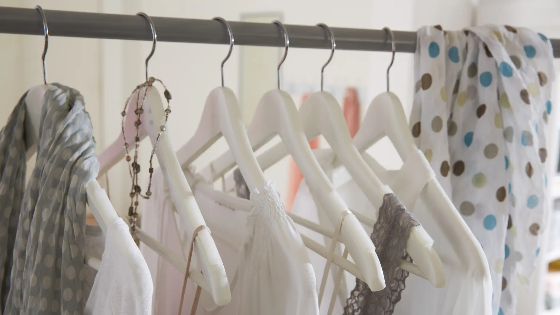A row of white clothes on white hangers is displayed on a metal rod. A necklace with beads hangs on one hanger, and a polka dot scarf drapes over the rod. The background is softly blurred.